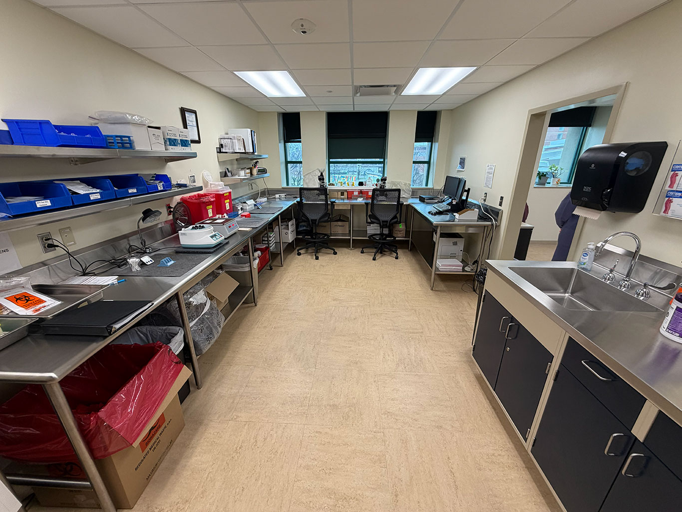 A clean, organized laboratory workroom with stainless steel counters and storage, bright overhead lighting, and a sink. Two chairs face a window.
