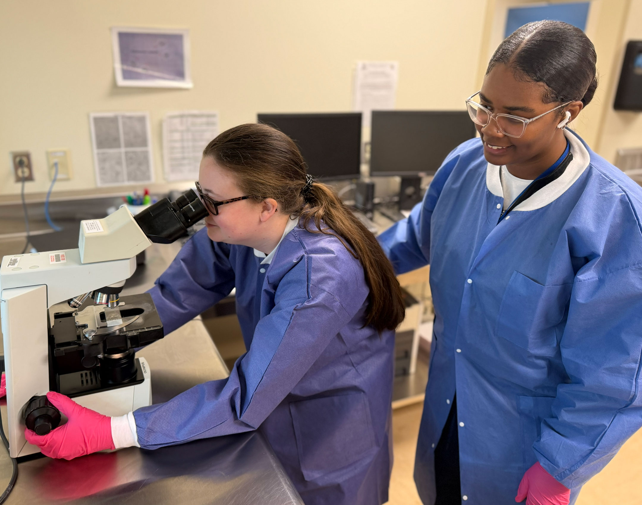 Two scientists in lab coats and safety goggles work in a laboratory. One peers through a microscope, while the other observes, both wearing pink gloves.