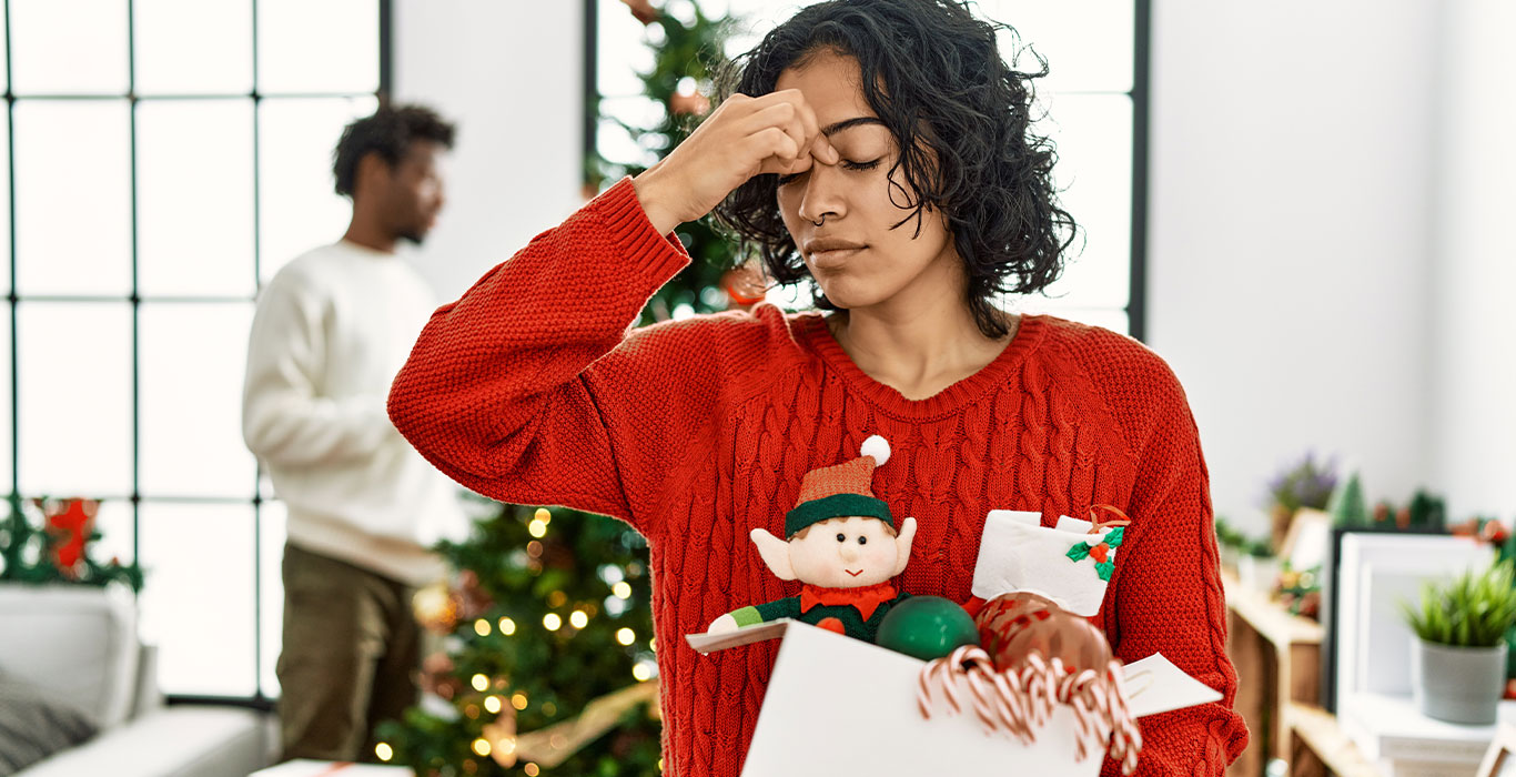 A woman in a red sweater looks stressed while holding a box of Christmas decorations, with a decorated tree and another person blurred in the background.