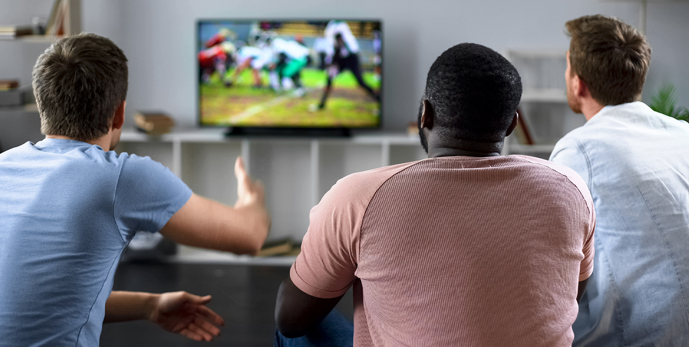 Three men sit on a couch, focused on a TV displaying a blurred football game. Their engaged posture suggests excitement and anticipation.