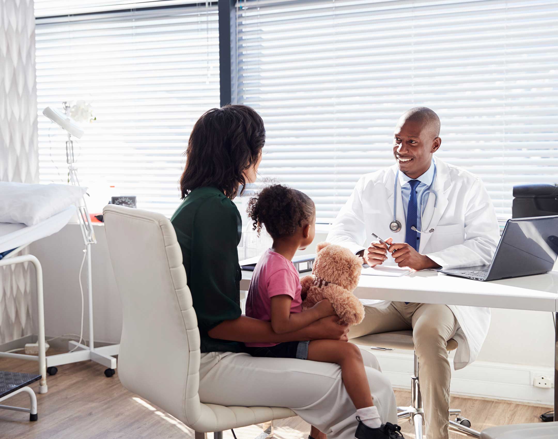 A doctor in a white coat smiles warmly while talking to a woman and her child, who holds a teddy bear, in a bright, modern medical office.