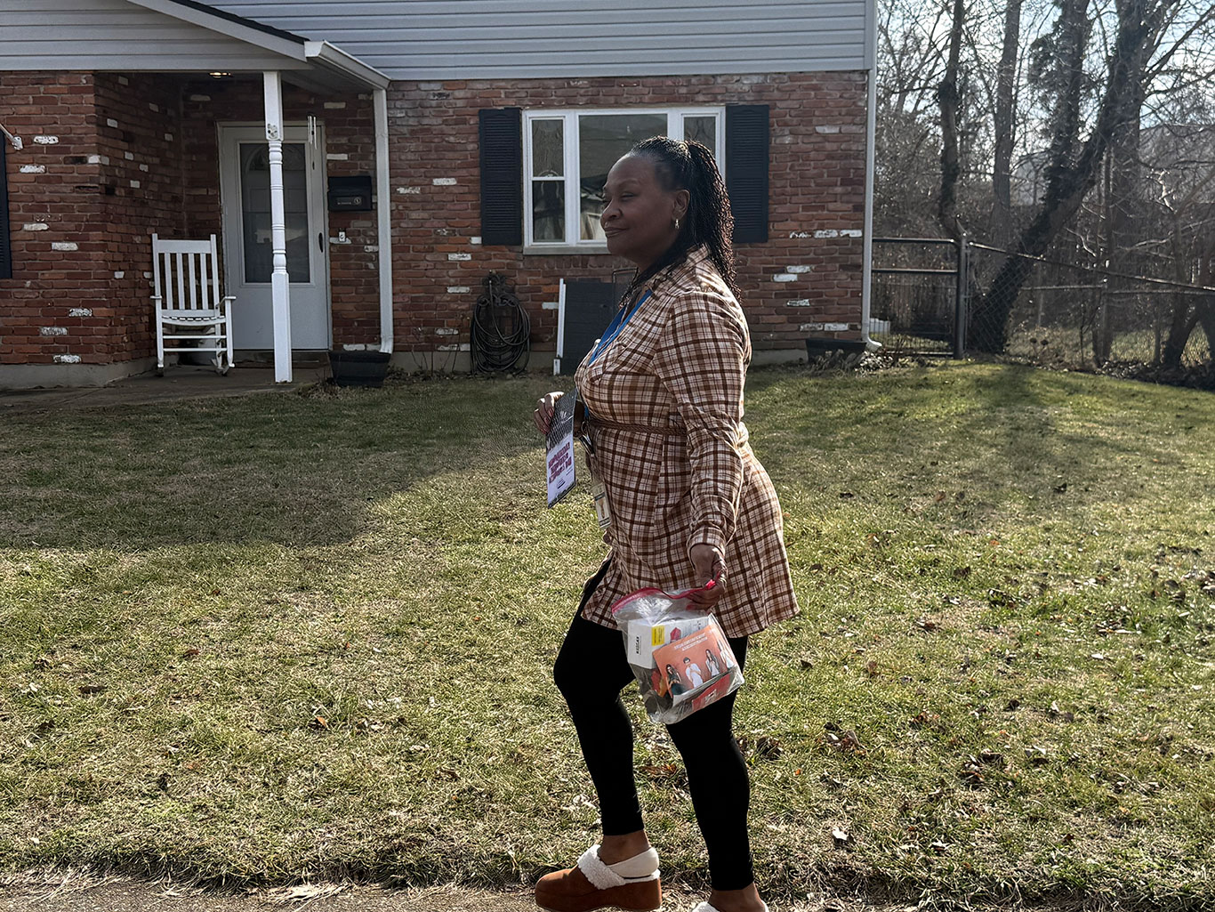 A woman walks on a lawn, holding a small clear bag and documents. She's in front of a red brick house with a white porch. The scene feels serene and sunny.