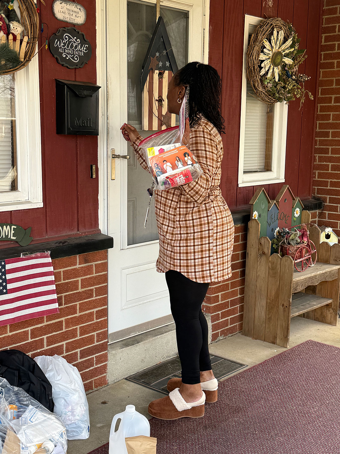 A person in a plaid shirt knocks on a red brick house door, holding a transparent bag with magazines. The porch has decorative wreaths and signs.
