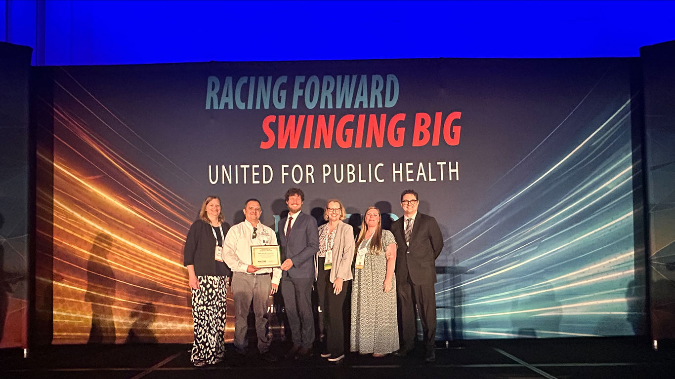 Six people stand on a stage in front of a banner reading Racing Forward, Swinging Big, United for Public Health, one holding an award plaque.